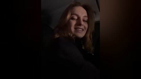 Media: A dimly-lit video of a smiling, fair-skinned woman with long, wavy blonde hair, wearing a dark jacket, taken from the passenger seat of a car.