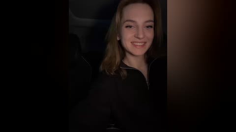 Media: Video of a smiling young woman with fair skin and shoulder-length light brown hair, wearing a black jacket, sitting in a dimly lit car.