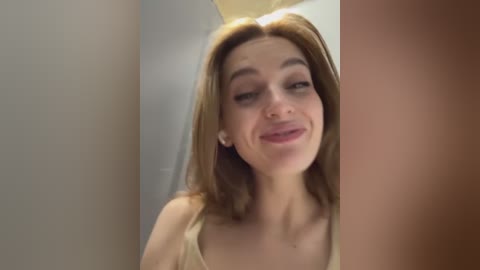 Media: Video of a young Caucasian woman with light brown hair, fair skin, and a slight smile. She wears a light-colored tank top, standing in a narrow, dimly lit hallway.