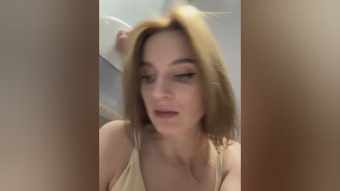 Media: A video of a light-skinned, fair-haired woman with a medium build, wearing a beige top, looking down with a slight frown. The blurred background suggests an indoor setting.