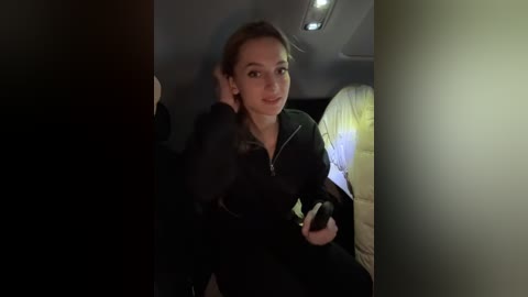 Media: Video of a young woman with light skin and brown hair in a black jacket, smiling and holding a smartphone, sitting in a dimly lit car.