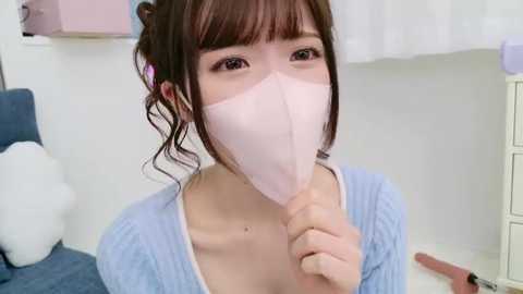 Media: Video of a young Asian woman with light skin, brown hair in a bun, wearing a pink face mask and light blue sweater, indoors with white walls and furniture.