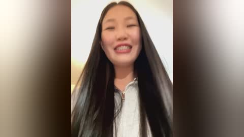 Media: A video of a smiling young Asian woman with long, straight black hair, wearing a light gray zip-up hoodie, set against a blurred, beige background.