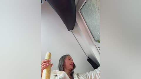 Media: A video of an Asian woman with long, straight, dark hair, holding a banana, wearing a white blouse, in a modern, minimalistic room with a metallic ceiling.