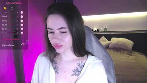 Media: Video of a young woman with light skin, long dark hair, and a white top, showcasing a tattoo on her chest, standing in a dimly lit bedroom with pink and purple lighting.