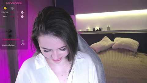 Media: Video of a young woman with long, dark hair, wearing a white blouse, sitting in a dimly lit bedroom with a bed and pillows in the background.