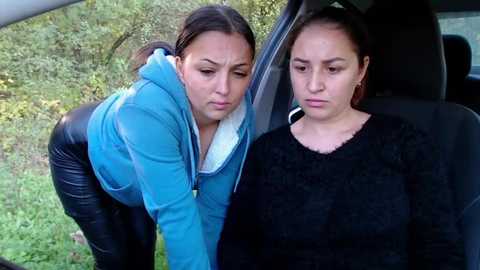 Media: Video of two women in a car: one with a blue hoodie, leaning in to comfort another in a black sweater, set against a blurred, green, outdoor background.