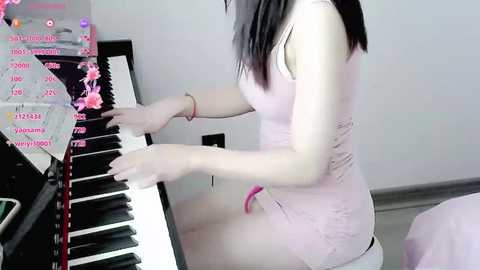 Media: Video of a slim, light-skinned woman with straight black hair, wearing a pink tank top and white shorts, playing a digital piano in a minimalist room.