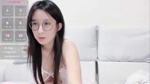 Media: Video of an East Asian woman with long black hair, wearing glasses and a light pink lace bra, sitting on a white bed in a clinical setting with a white wall and a \"Lounger\" poster.