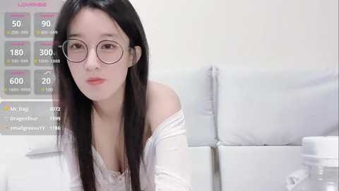 Media: Video of a young Asian woman with long black hair, wearing glasses, and a white off-shoulder top, sitting on a white couch, with a digital health tracker display in the background.