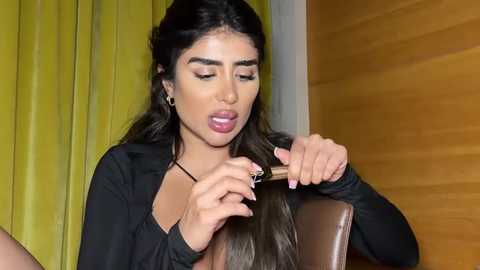 Media: Video of a Latina woman with long, wavy hair, medium skin tone, and full lips, styling her hair with a comb while sitting in a chair, wearing a black top, against a yellow curtain and wooden wall.