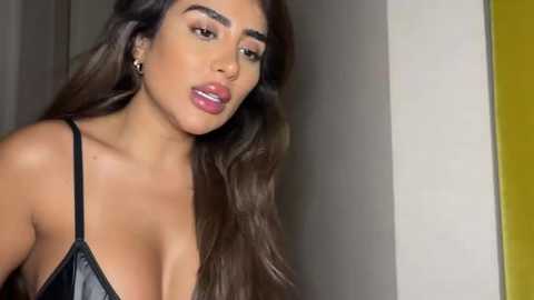 Media: Video of a woman with long, dark hair, wearing a black leather bra, standing in a room with a yellow and white wall. She has full lips and a confident expression.