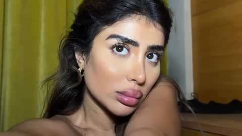 Media: Video of a young woman with olive skin, long dark hair, full lips, and a nose ring, wearing a gold hoop earring, in a bedroom with a yellow curtain and wooden door.