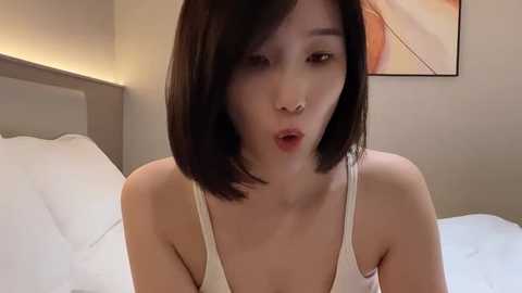 Media: Video of an Asian woman with straight, shoulder-length black hair, wearing a white tank top, making an exaggerated, surprised face in a modern bedroom with white sheets and a beige headboard.