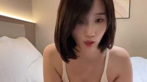 Media: A video of an East Asian woman with straight, shoulder-length black hair, light skin, and a slender physique, wearing a white tank top, sitting on a bed with white sheets in a modern, minimalist bedroom.