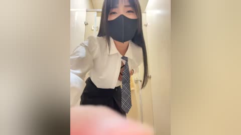 Media: Video of an Asian woman with long black hair and a white shirt, black mask, and blue tie, standing in a dimly lit bathroom, looking down.