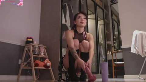 Media: Video of a slim, tattooed, Asian woman with dark hair in a black leotard, squatting with a large dildo, in a cluttered, dimly lit bedroom with a purple chair and shelves.