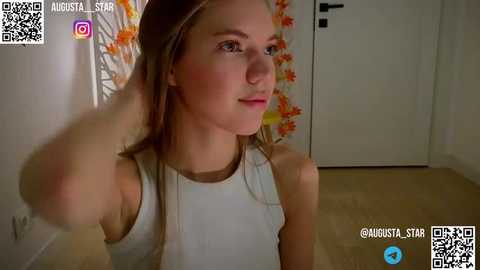 Media: Video of a young Caucasian woman with light skin and brown hair, wearing a white tank top, standing in a room with a floral curtain and wooden floor.