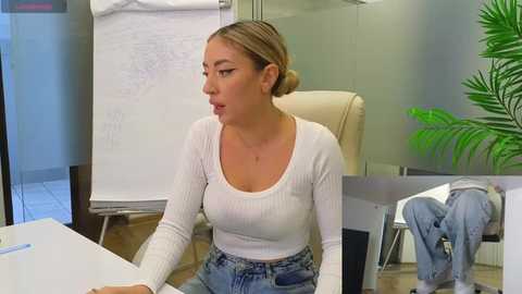 Media: Video of a light-skinned woman with blonde hair in a bun, wearing a white ribbed top and high-waisted jeans, sitting at a desk in an office with a whiteboard, plants, and a chair.