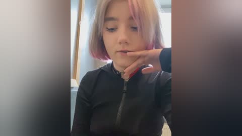 Media: Video of a young woman with shoulder-length, pastel pink and blonde hair, wearing a black zip-up hoodie. She appears to be fiddling with her mouth. The background is blurred, with a hint of a window.