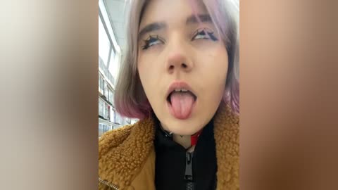 Media: Video of a young woman with pale skin, pink hair, and a tongue out expression. She wears a brown fuzzy coat and a black hoodie, standing in a bookstore with shelves in the background.