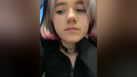 Media: Video of a young woman with shoulder-length, pastel-colored hair, wearing a black zip-up hoodie and a black choker with a heart pendant. She has a neutral expression, with a soft-focus background.