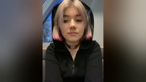 Media: Video of a young woman with pastel pink and white hair, wearing a black hoodie and choker, seated indoors by a window, looking down, blurred background.