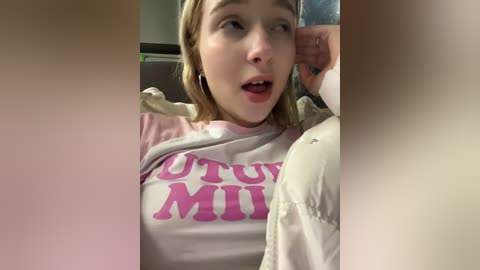 Media: Video of a young, fair-skinned woman with blonde hair, wearing a light pink \"Future Mi\" sweatshirt, captured indoors, looking surprised.