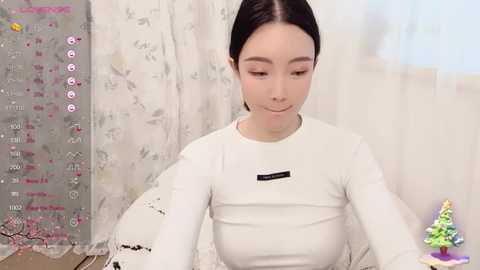 Media: Video of an Asian woman with fair skin and long black hair, wearing a white long-sleeve shirt, sitting in front of a wall with a patterned calendar. The background has a decorative Christmas tree.