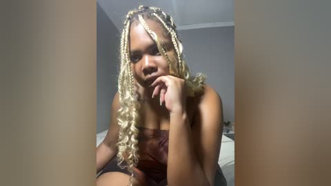 Media: Video of a young Black woman with light brown skin, styled with long, blonde braids, wearing a dark top, resting her chin on her hand, indoors with neutral walls.