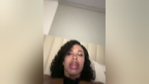 Media: A blurry video of a woman with curly black hair, closed eyes, and open mouth, lying on a beige bed in a minimalistic room with cream walls.