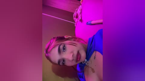 Media: Video of a young woman with fair skin and wet, pink-dyed hair, wearing a blue dress, and a silver necklace, taken in a dimly lit room with pink and purple lights.