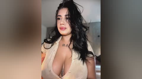 Media: Video of a young, light-skinned woman with long, wavy black hair in a high ponytail, wearing a low-cut, light-colored blouse that reveals ample cleavage. She has a tattoo on her collarbone. Background is out of focus, likely indoors.