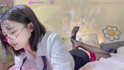 Media: A video of an Asian woman with short black hair, wearing glasses, lying on a bed, dressed in a white shirt, black skirt, and black stockings, with a digital filter overlay.