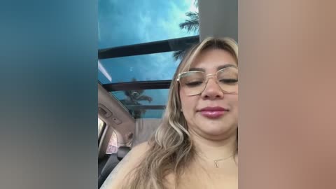 Media: Video of a smiling, light-skinned woman with long, wavy blonde hair and glasses, wearing a necklace, sitting in a car with a view of a clear blue sky and palm trees through the window.