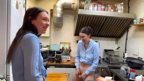 Media: Video of two women, one wearing a blue shirt and the other nude, in a kitchen with stainless steel appliances and cooking utensils.