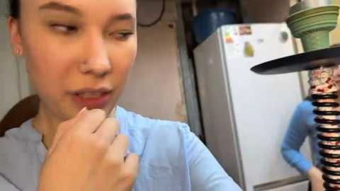 Media: Video of a young woman with fair skin and short dark hair, wearing a light blue shirt, in a kitchen, holding a spoon near a white fridge with magnetic decorations.