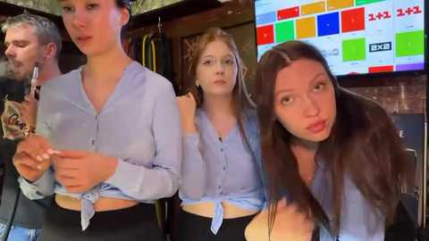 Media: Video of three women in light blue tops, one with a tie, sitting close, smiling, in a brick-walled room with a TV screen displaying colorful graphics.