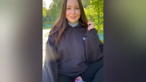 Media: Video of a smiling woman with long brown hair, wearing a black hoodie, standing outdoors by a lake, surrounded by greenery.