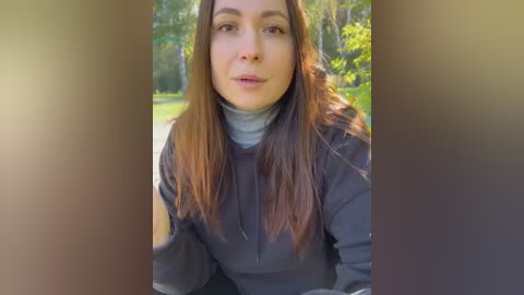 Media: A video of a fair-skinned woman with long brown hair, wearing a dark jacket and gray turtleneck, standing outdoors in a sunlit park with green trees and a paved path in the background.