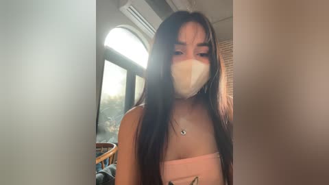 Media: Video of an Asian woman with long, straight black hair, wearing a pink strapless dress and a white face mask, standing indoors near a window with greenery visible outside.