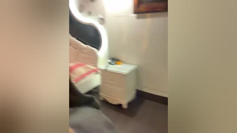 Media: A blurry video of a small, dimly lit room with a white bed, black chair, and beige walls. A white dresser with a single drawer is visible, and a framed picture hangs on the wall.