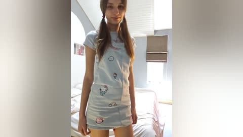 Media: A young, slender, light-skinned woman with long brown hair in pigtails wears a light blue, short, t-shirt dress adorned with Hello Kitty designs. She stands in a bright, modern bedroom with a bed, window, and soft lighting.