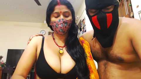 Media: Video of a woman with long black hair and fair skin, wearing a black bra, face paint, and red necklace, posing with a shirtless man in a black mask.