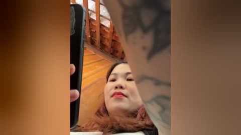 Media: A video of a smiling, fair-skinned woman with long, curly red hair, wearing a black top, taking a selfie with a black phone. The background features a wooden structure and blurred text.