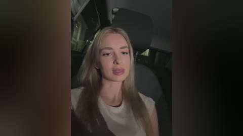 Media: Video of a blonde woman with straight hair, light skin, wearing a white blouse, sitting in a dimly lit car interior.