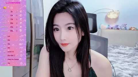 Media: A video of an East Asian woman with long black hair and fair skin, wearing a green top, sitting in a modern bedroom with a purple calendar, plush toys, and a bed.