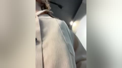 Media: Video of a person wearing a light-colored, textured shirt, captured from a low angle, with blurred background featuring a ceiling light.