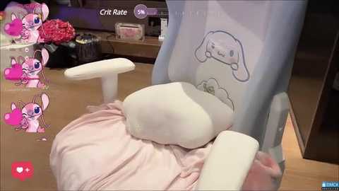 Media: Video of a plush pink and white pillow on a gaming chair, featuring an animated character with a sad expression. Background includes a wooden desk with a lamp and red flowers.