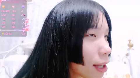 Media: A video of a young East Asian woman with long black hair, straight bangs, and a light complexion. She is smiling, and the background is a blurred bedroom with white furniture.